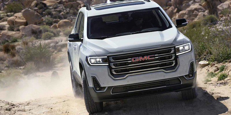 A white GMC Acadia driving off-road through a rocky desert terrain.