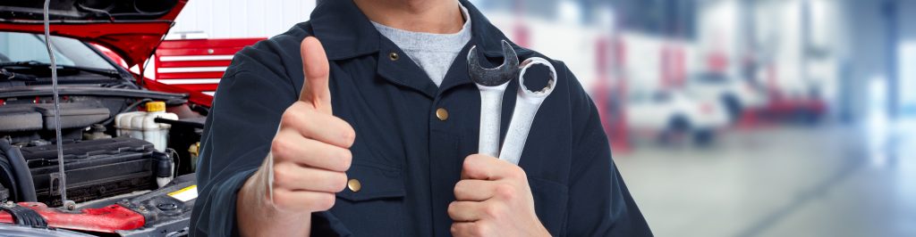 A mechanic giving a thumbs up while holding two wrenches in front of a car engine.